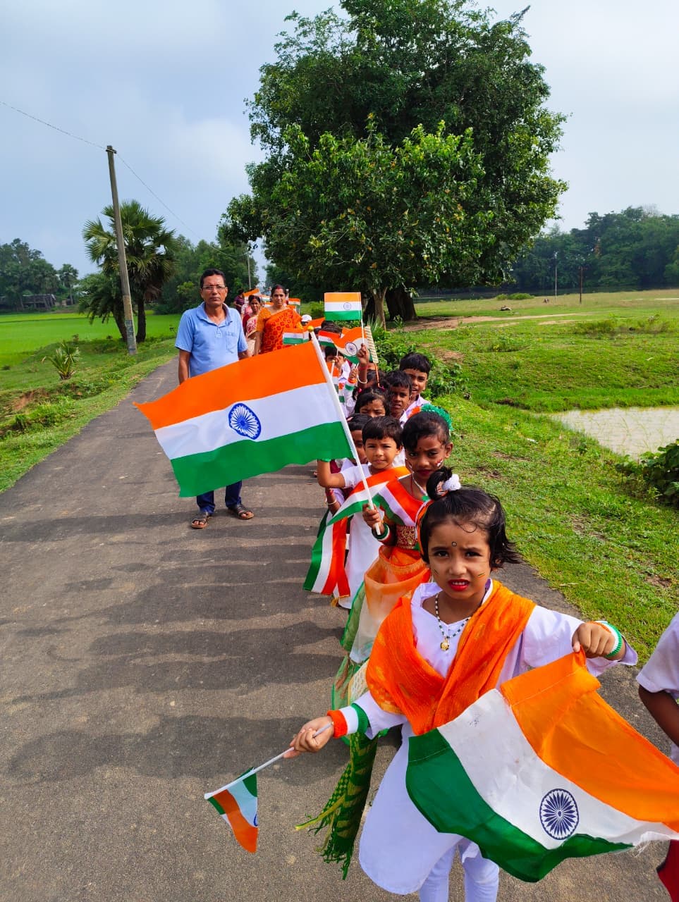 Children waving Indian flags as they walk along a village road between green fields