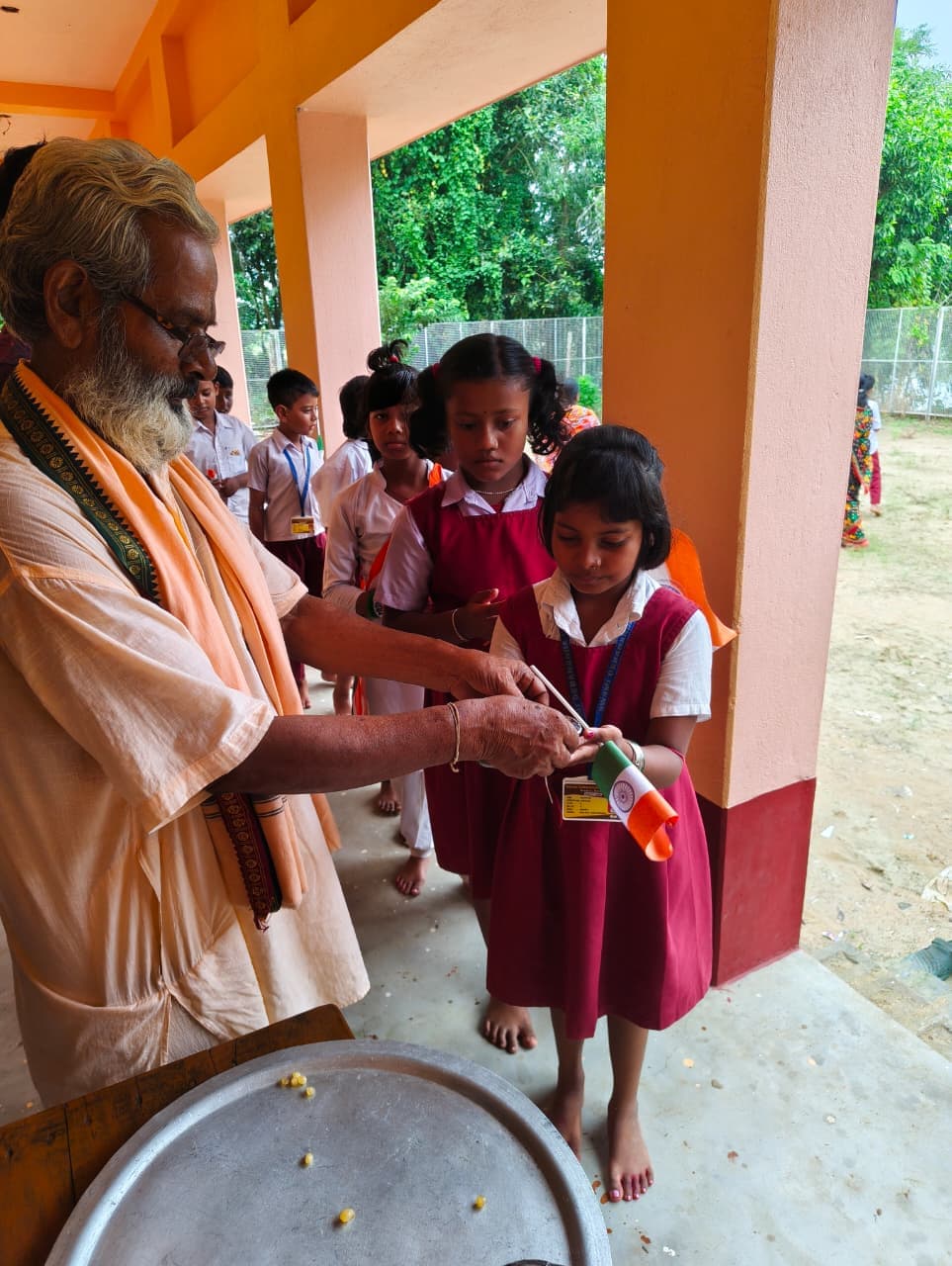 A man in saffron handing a small flag to schoolgirls in maroon uniforms on a veranda