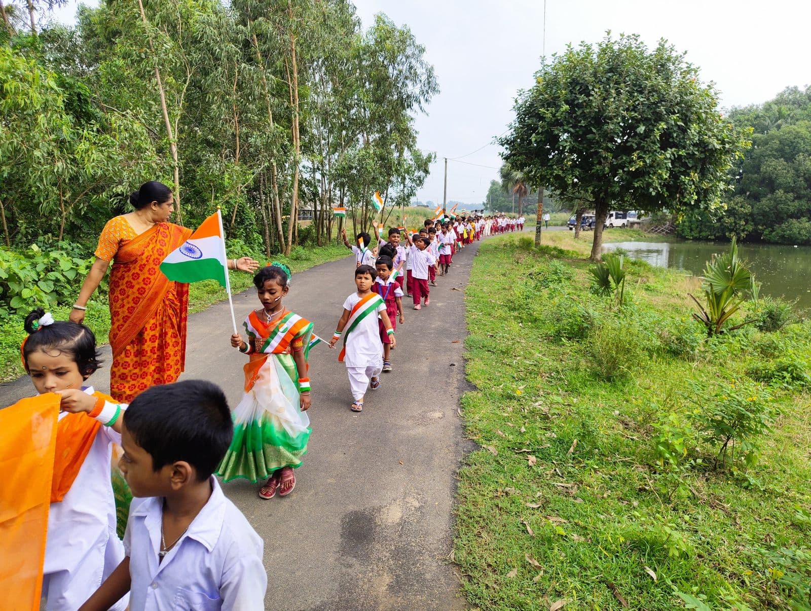A line of children carrying Indian flags walking along a road beside a pond, led by a woman in an orange sari