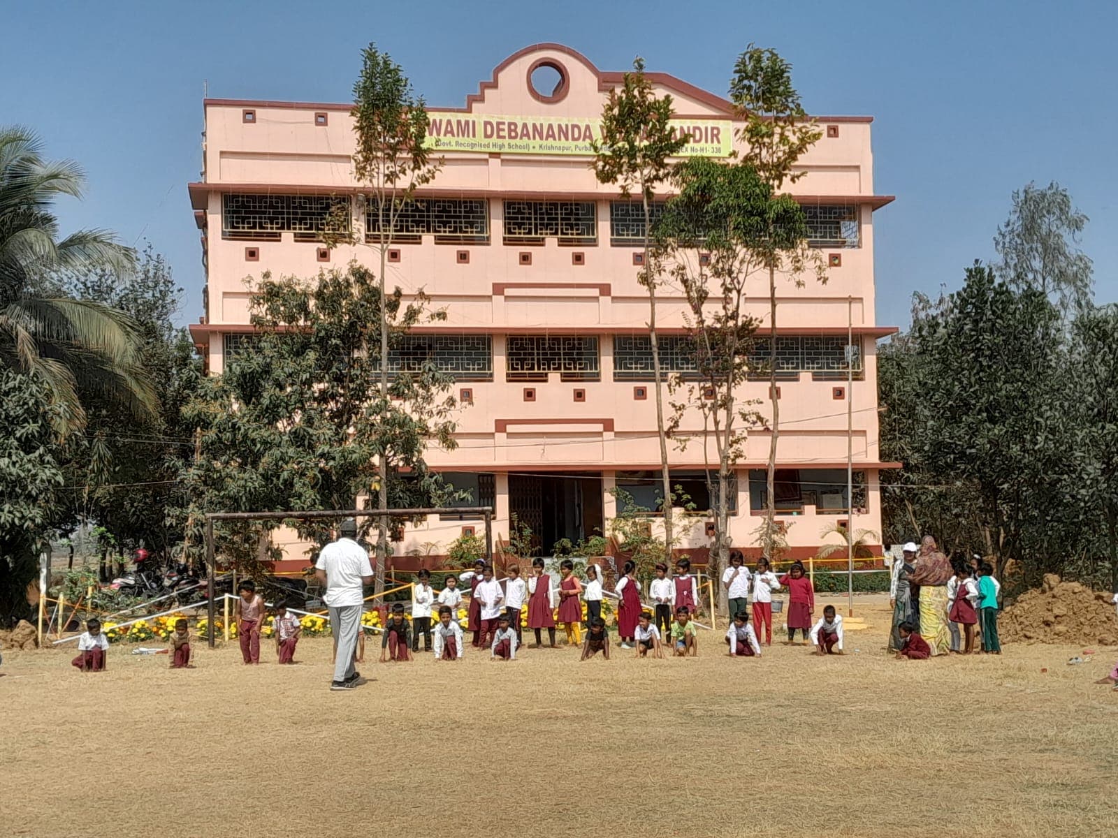 The pink multi-storey school building across an open field, with students lined up in front