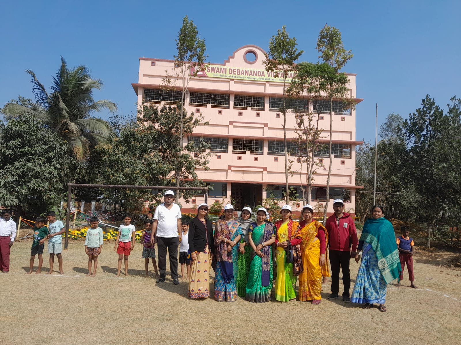 A group of teachers and children standing on the field in front of the pink multi-storey school building