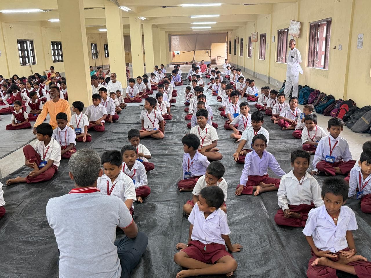 Rows of students in uniform seated cross-legged on mats in a long hall