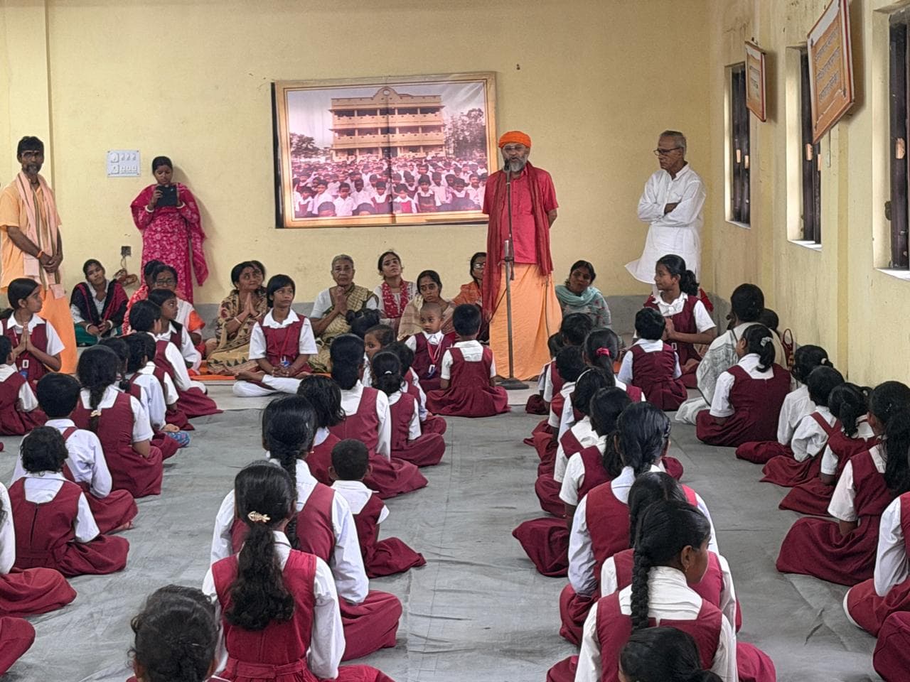 Students in maroon uniforms seated on the floor of a hall, facing a man in saffron standing before a framed photograph of the school