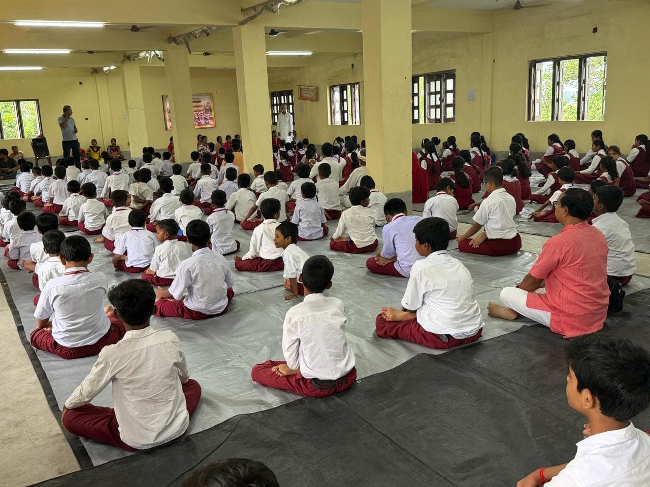 Students in white shirts seated in rows on mats in a large pillared hall