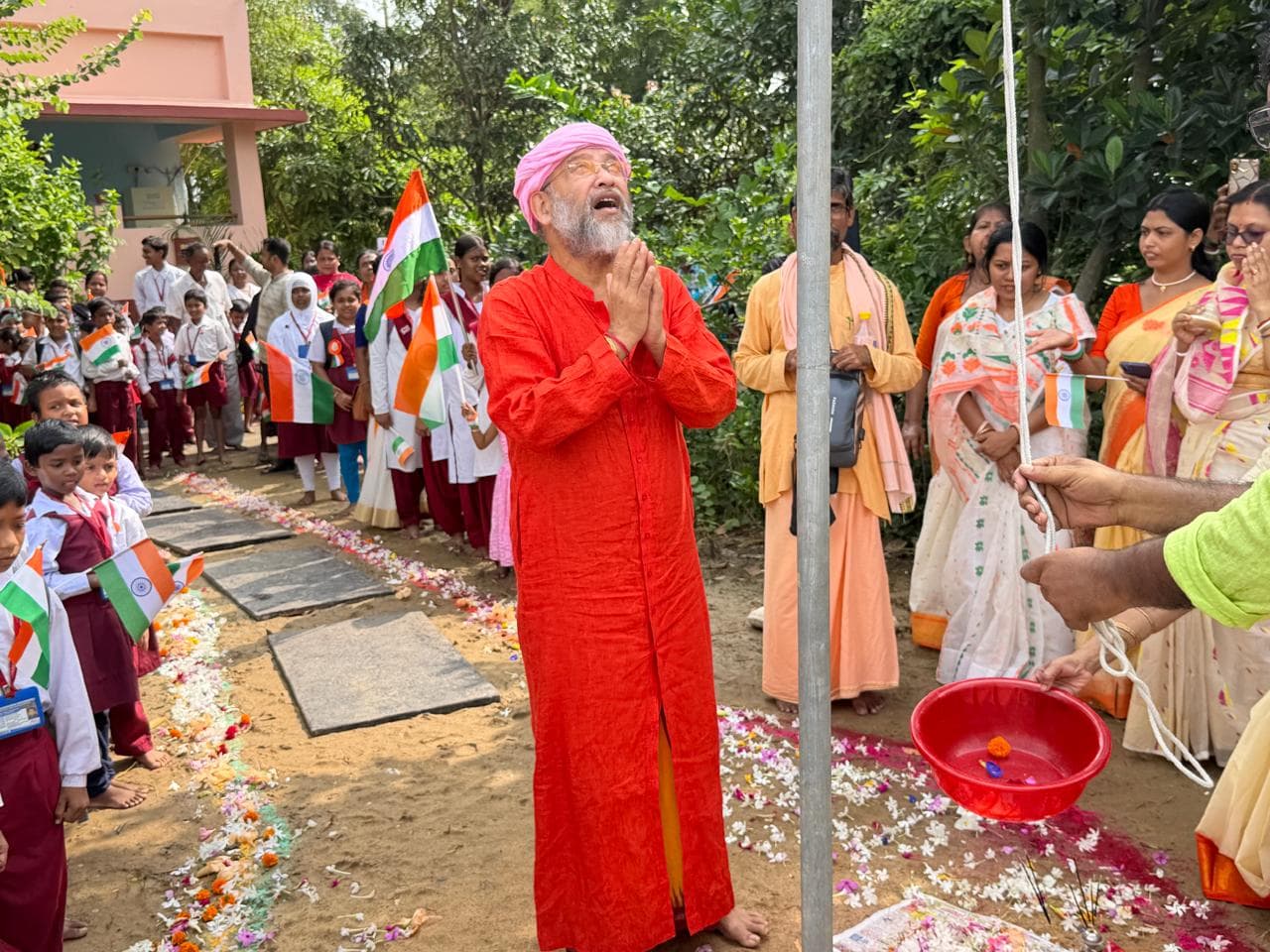 A man in a red robe and pink turban with hands joined beneath a flagpole, surrounded by students holding small flags