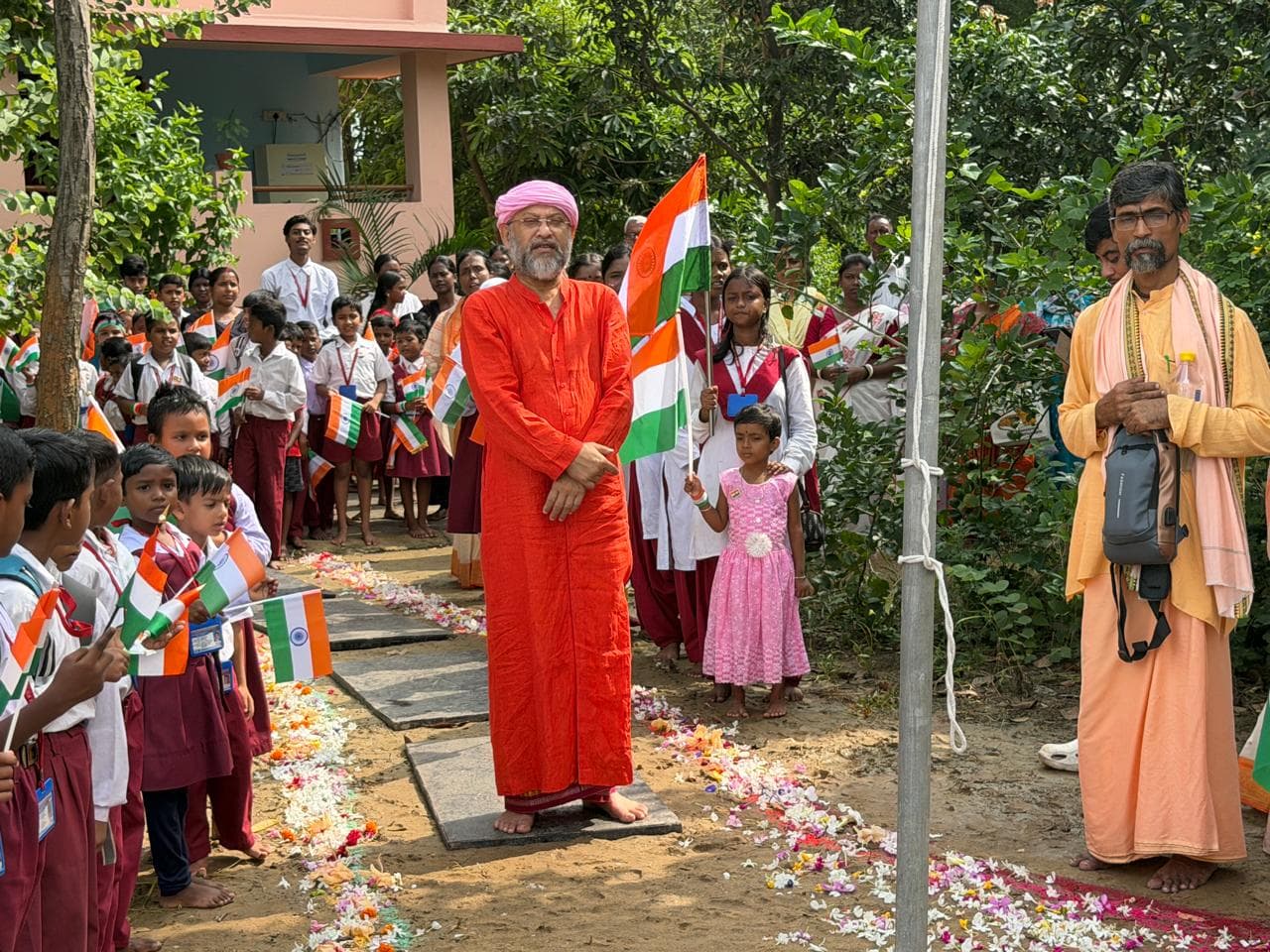 A man in a red robe standing by a flagpole, with students holding small flags and a man in saffron beside him