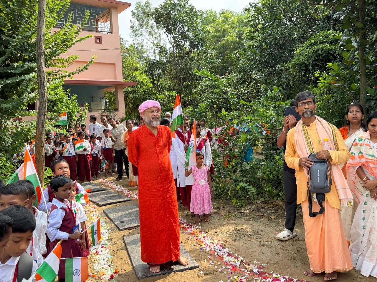 A man in a red robe beneath a flagpole on a path strewn with petals, with students holding flags gathered around