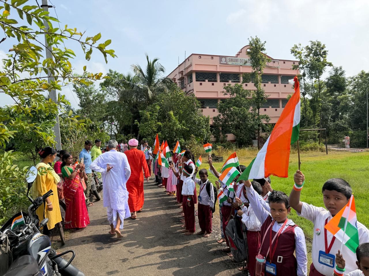 Students holding Indian flags lining a path as a procession passes, with the pink school building beyond the trees