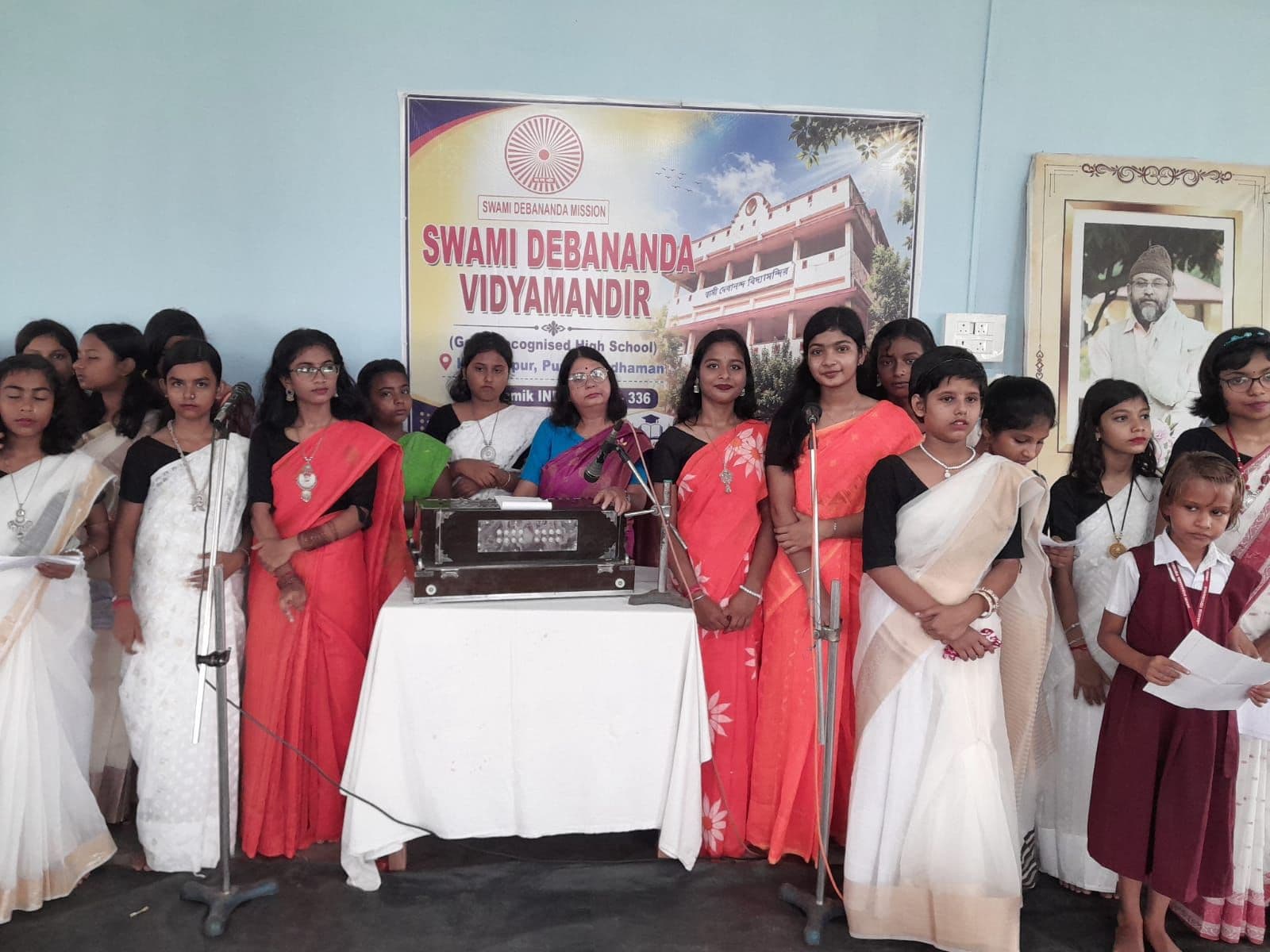 Girls in white and red saris standing behind a harmonium and microphones, in front of a banner of the school