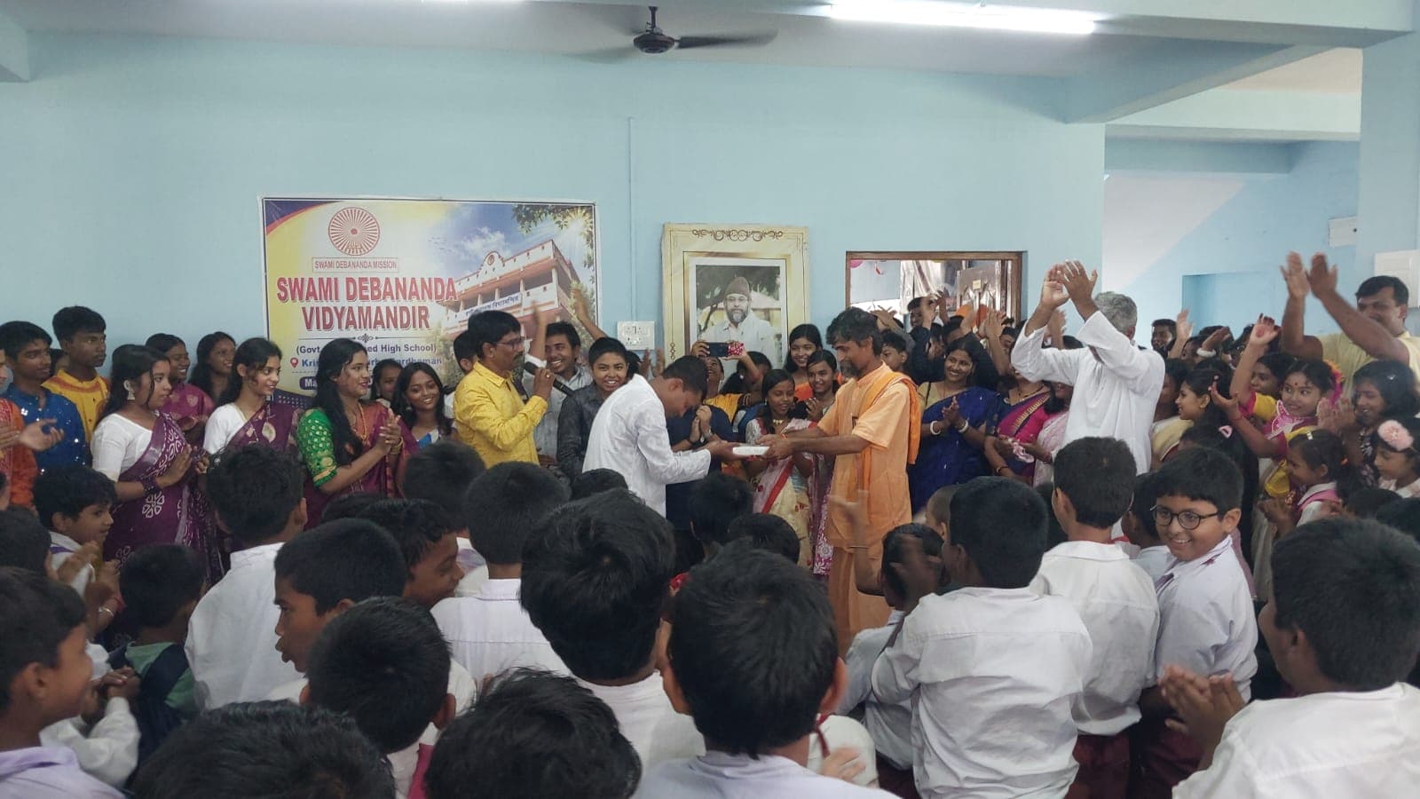 Students gathered in a hall, some raising their hands, in front of a banner of the school