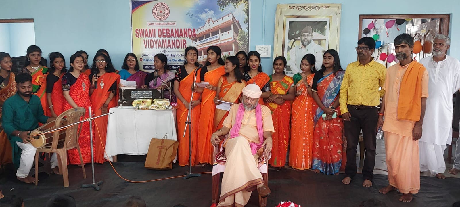 A group in orange saris and saffron clothing on a stage in front of a banner of the school, with a harmonium and microphones