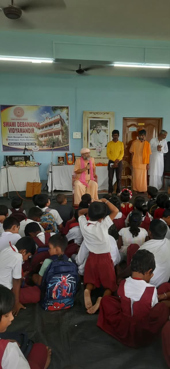 Students seated on the floor of a hall facing a table and a banner of the school