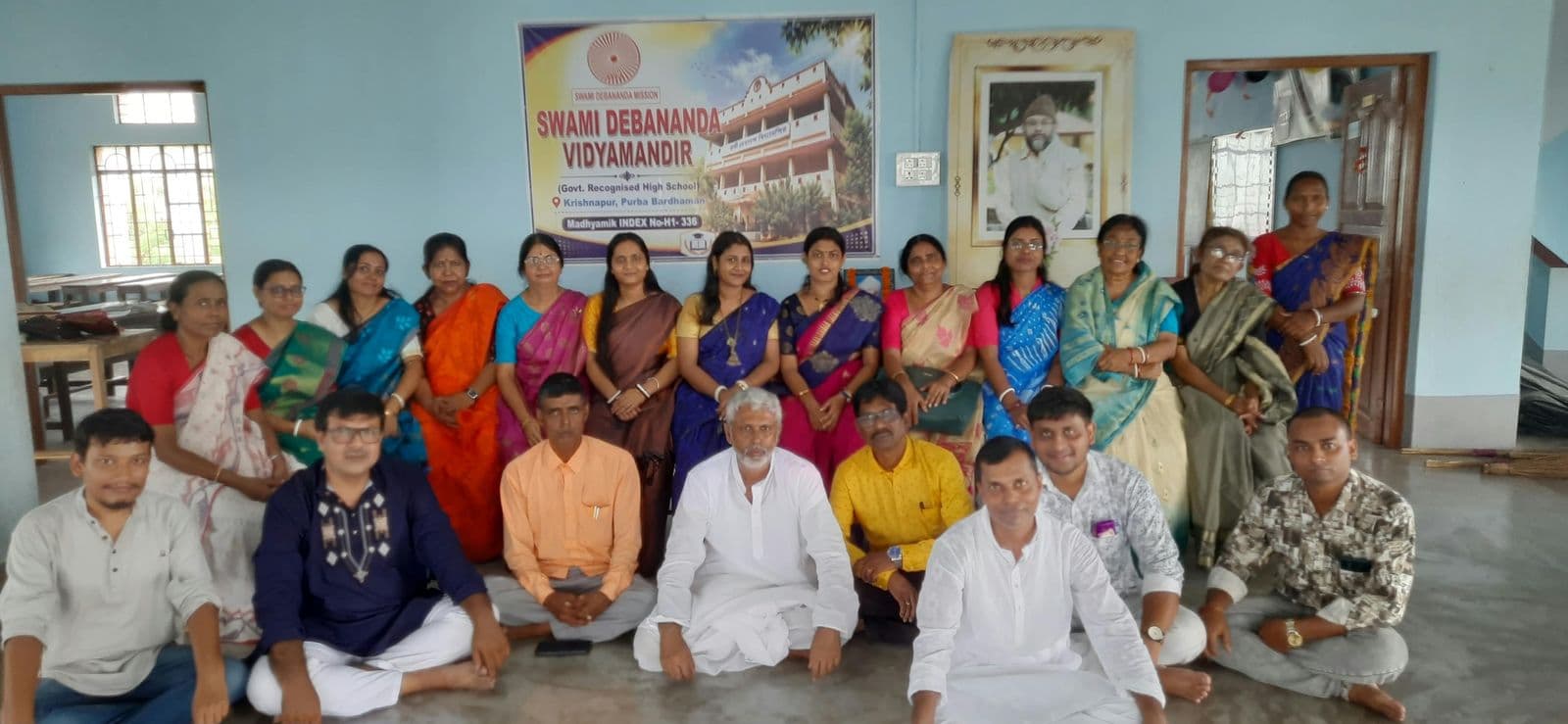 Teachers and staff seated and standing in rows in front of a banner of the school