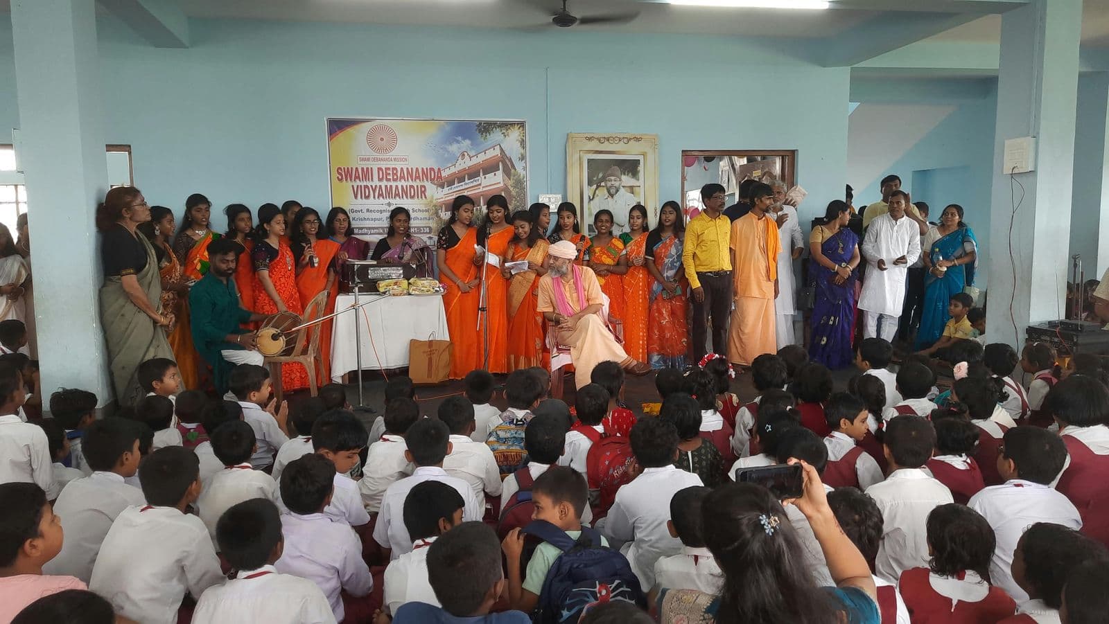 Students seated in a hall watching a group perform on a stage beneath a banner of the school