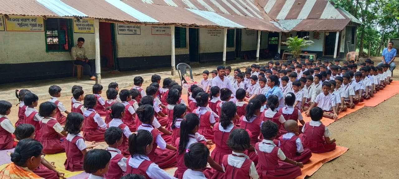 Students in red and white uniforms seated in rows on mats in front of a long building with a tin roof