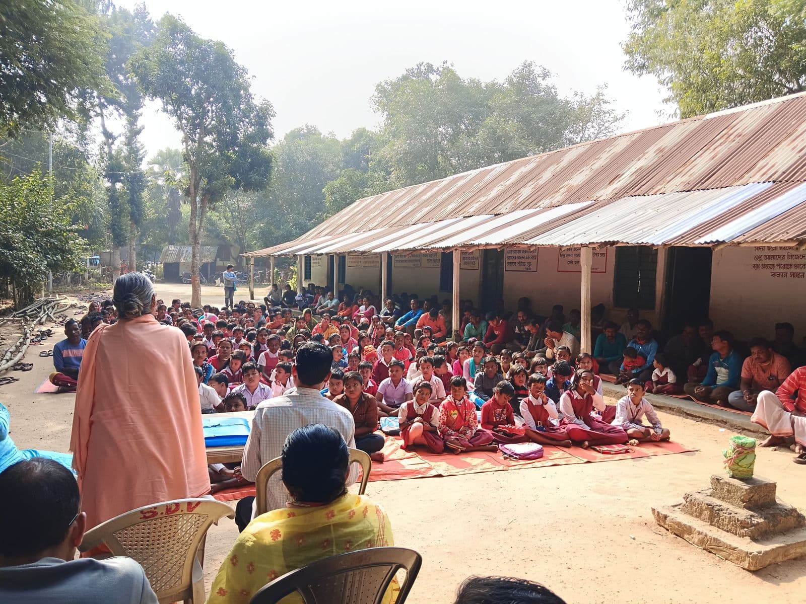 Students seated on mats in a yard before a long tin-roofed building, with a man in saffron facing them
