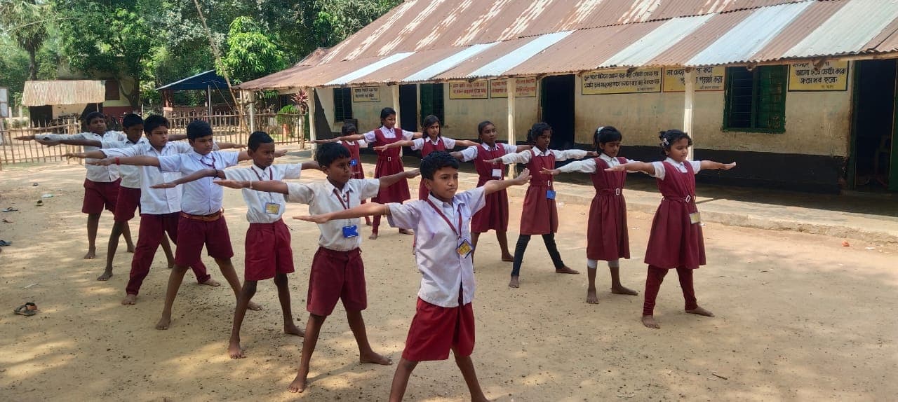 Students in uniform doing stretching exercises with arms outstretched in front of a tin-roofed school building