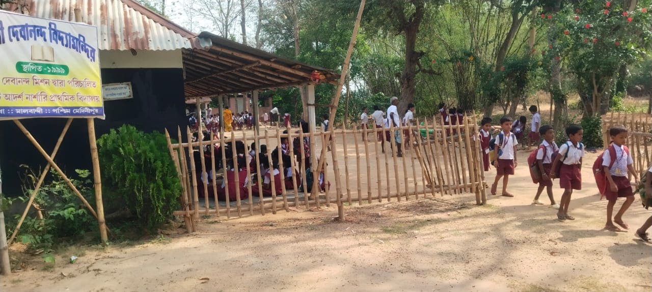 Children in uniform running across a sandy yard beside a bamboo fence and a signboard lettered in Bengali