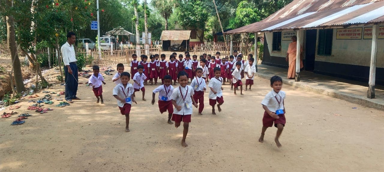 Children in uniform running across a sandy yard towards the camera, in front of a tin-roofed building