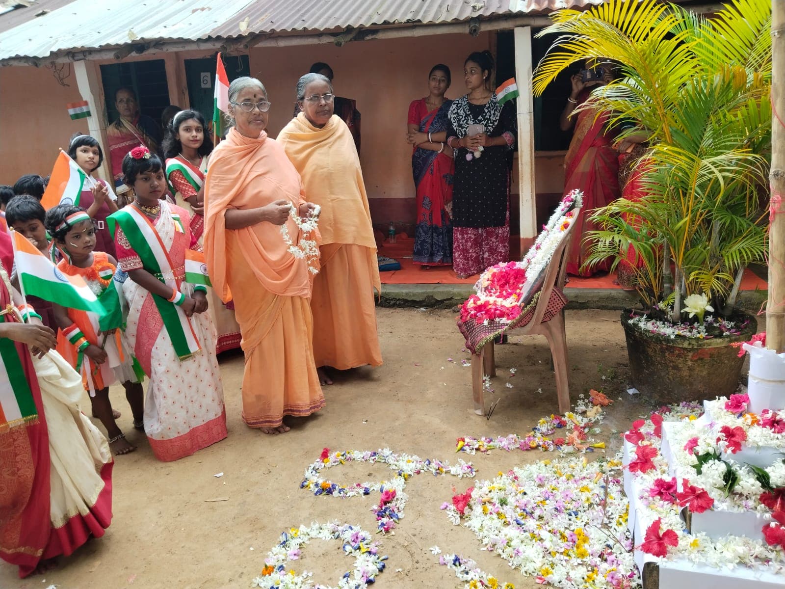 Two women in saffron standing beside children holding flags, with flower petals arranged on the ground