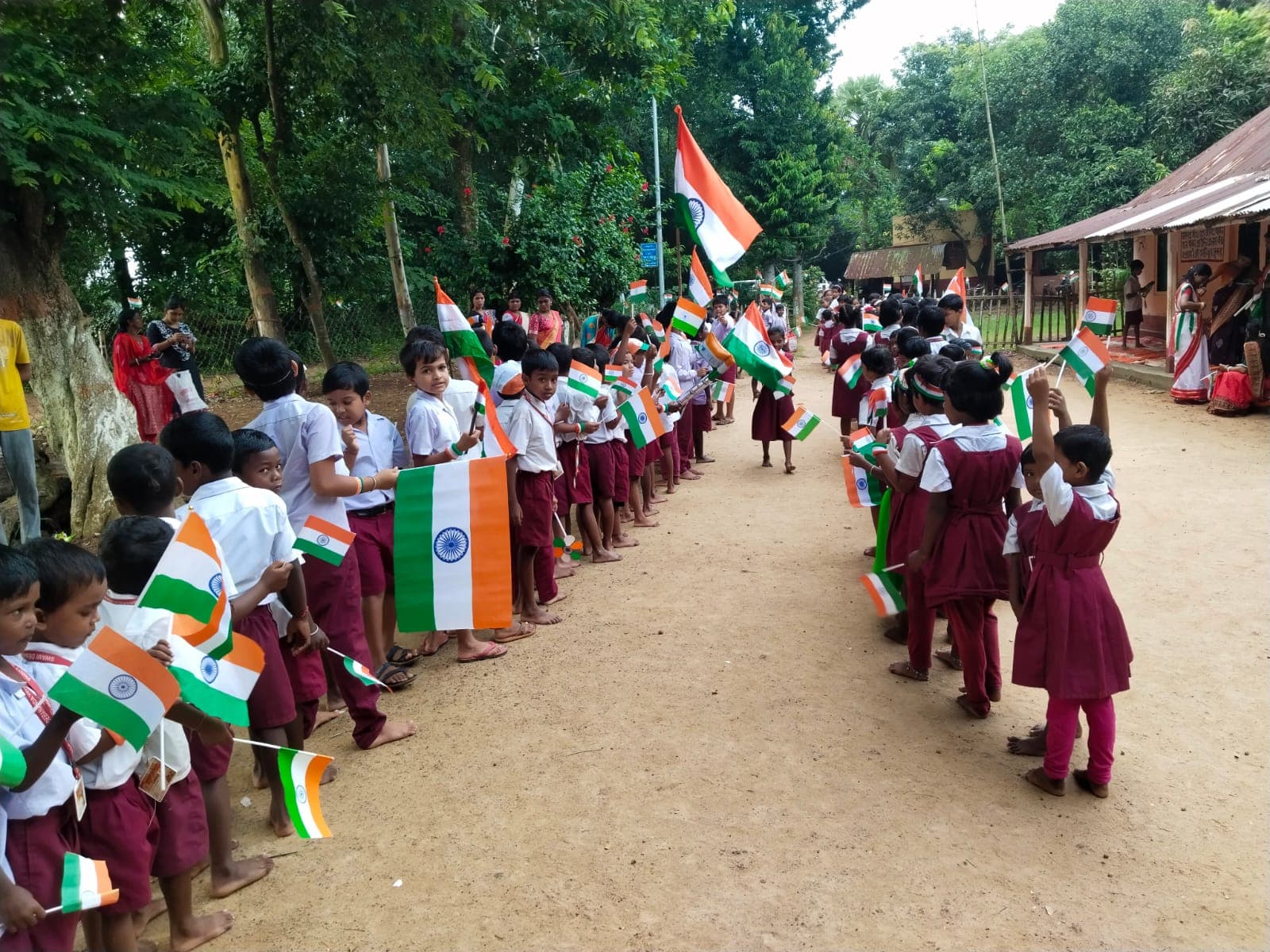 Two lines of students holding Indian flags along a sandy path through trees