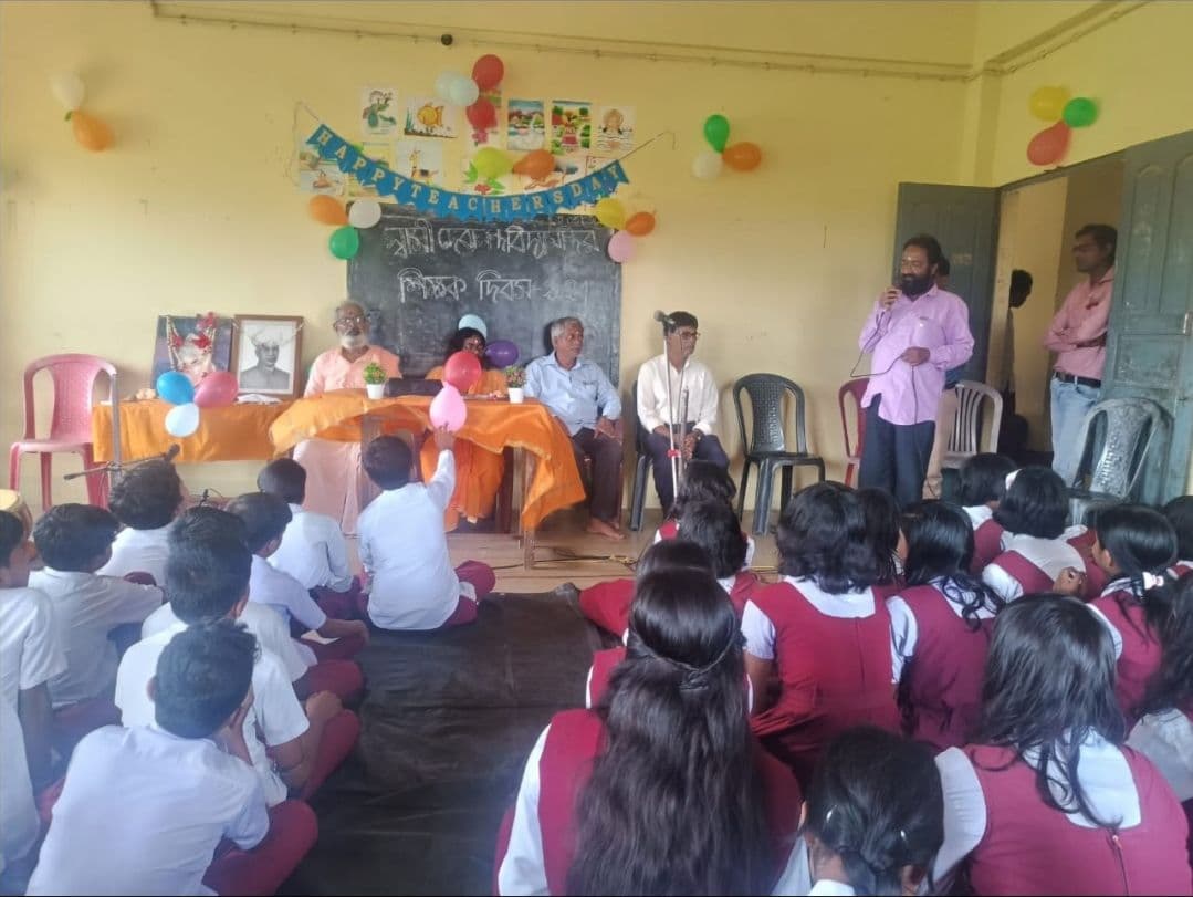 Students seated on the floor of a classroom decorated with balloons and paper bunting, facing a row of seated guests