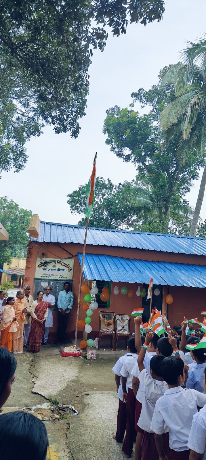 Students with flags gathered in front of a single-storey school building with a blue roof, under tall trees