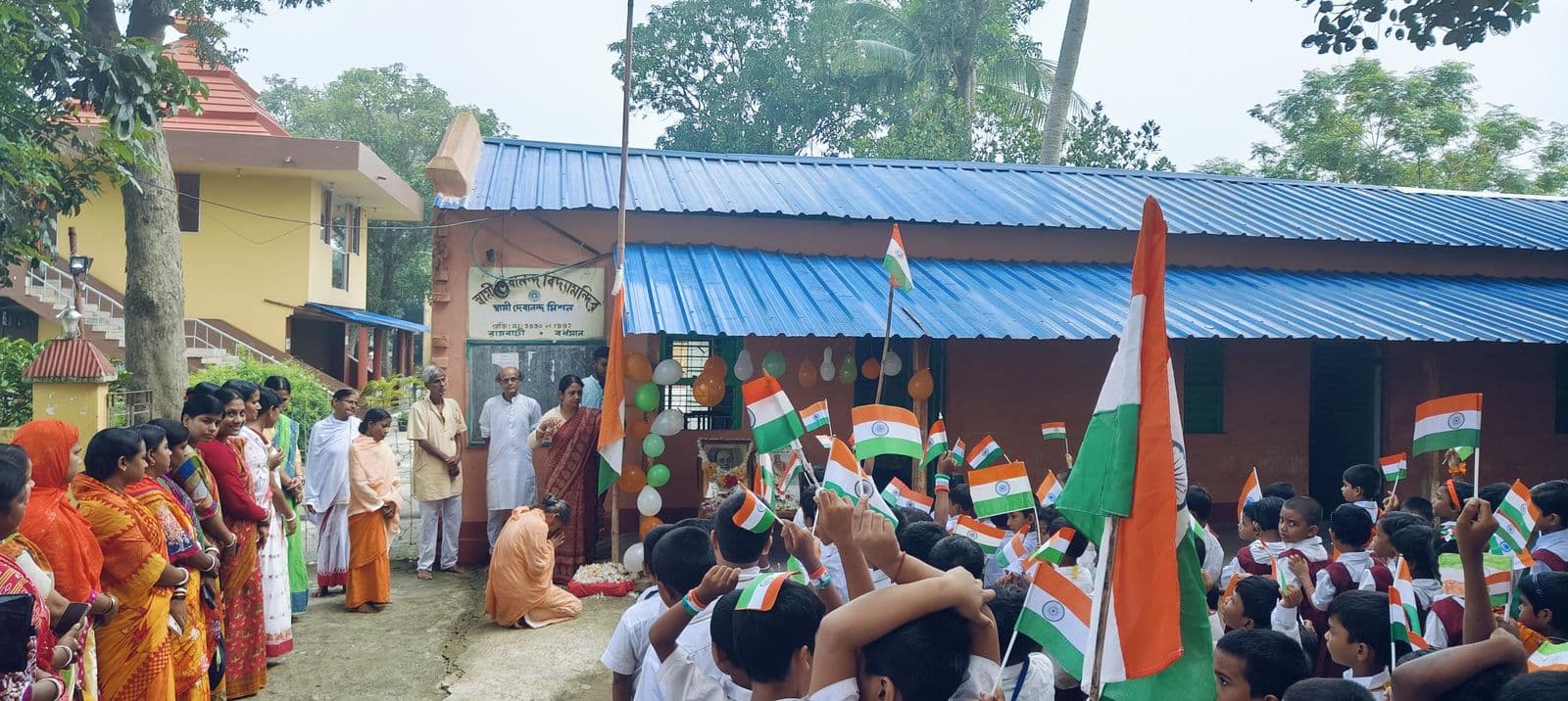 A gathering of students and adults holding Indian flags in front of a blue-roofed school building