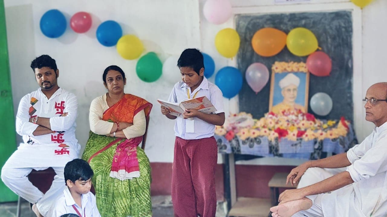 A student reading aloud beside two seated adults, in front of a garlanded portrait and a wall of balloons