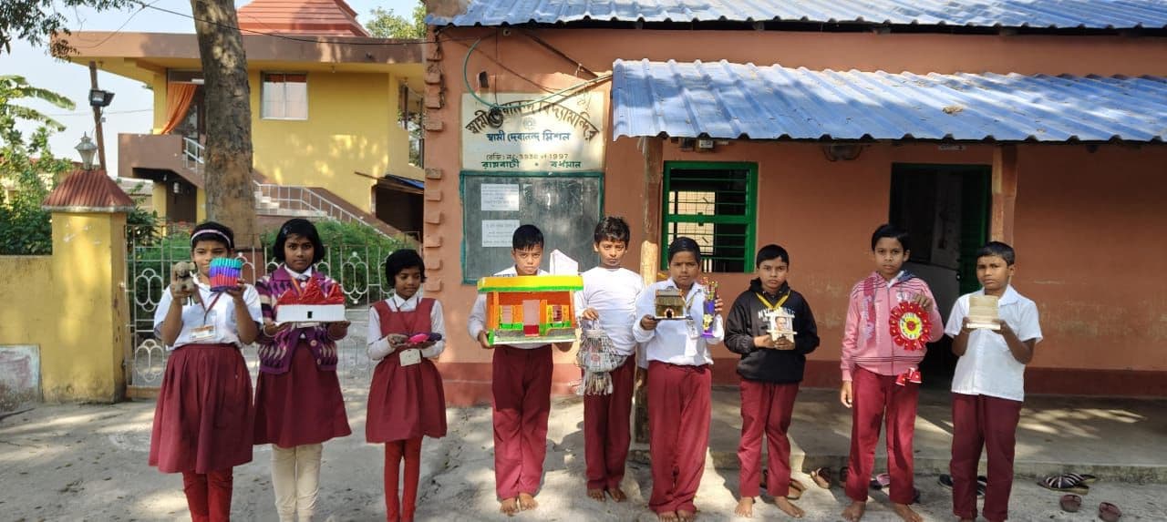 A line of students in uniform holding up small handmade models in front of a school building