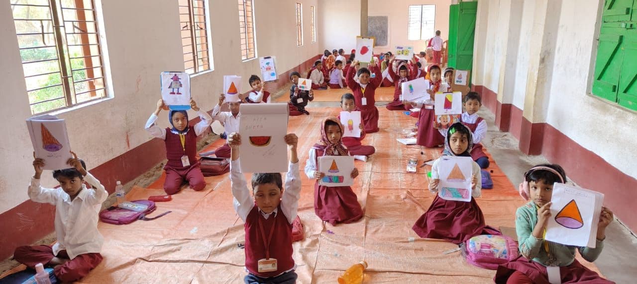 Students seated in rows on the floor of a classroom, holding up drawings of shapes