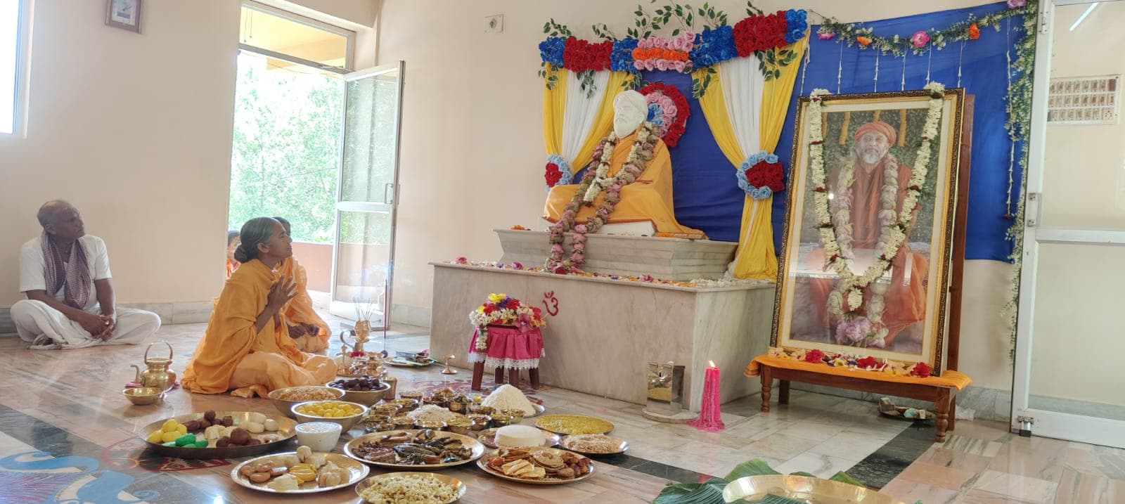 An altar with a seated statue and a garlanded framed portrait, with plates of food offerings on the floor and two people seated nearby
